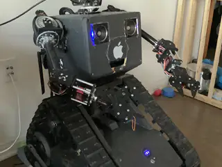 Black tracked robot with blue-lit camera eyes and articulated arms, seen from the front corner.