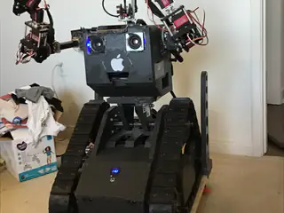 Black tracked robot faces forward with both articulated arms raised and blue camera lights glowing.