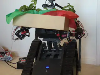 R.O.B. supports a broad planter filled with green foliage while its camera head turns.