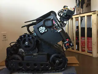 Side profile of a compact black tracked robot with exposed arm mechanisms and electronics.