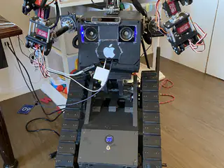 Front view of a black tracked humanoid robot with raised arms, camera eyes, and exposed wiring indoors.