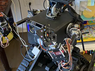 Three-quarter close view of the tracked robot showing its camera face, shoulder mechanisms, arm wiring, and tank treads.