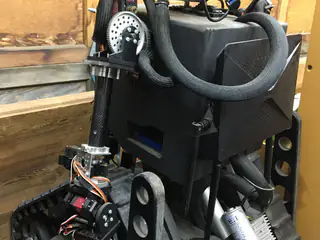 Rear three-quarter view of the black enclosure, curved braided conduit, arm actuator, and tank tread.