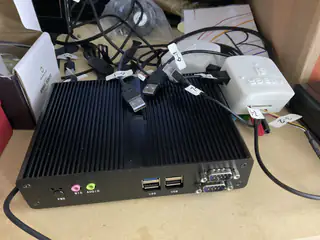 Fanless black mini computer with labeled USB cables and serial ports on a cluttered workbench.