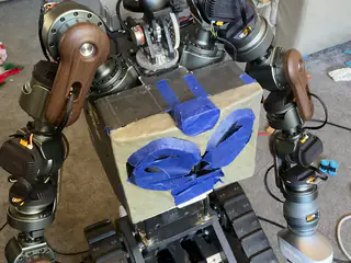 Tracked robot with articulated arms and a temporary chest enclosure decorated with blue painter's tape.