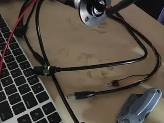 USB and power cables surround a flanged rotary connector resting beside a laptop and metal bracket.
