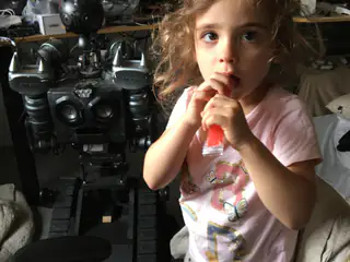 Young child holding a snack beside a black humanoid robot in a cluttered living room.