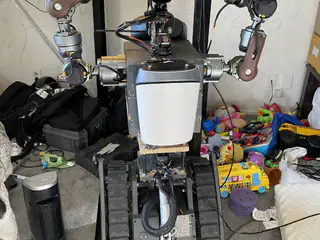 Front view of the tracked robot with a white chest panel and both articulated arms raised among toys.