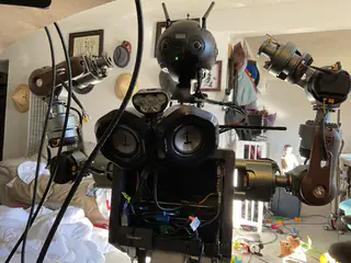 Close frontal view of the robot's spherical camera head, glowing green light, twin speakers, and raised arms.