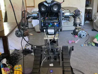 Full frontal robot with twin speakers, exposed chest wiring, extended arms, and a tracked base.