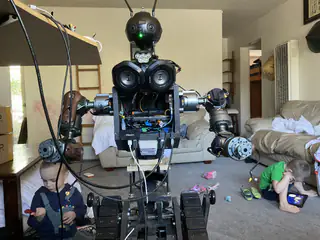 Wide living-room view of the tracked robot, with children partially visible behind its extended arms.