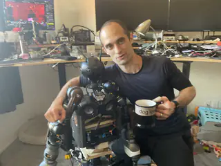 Seated man holding a mug beside the partially assembled humanoid robot in a cluttered workshop.