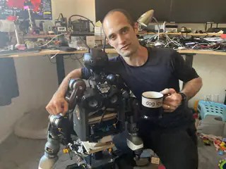 Closer framing of a man holding a mug beside the robot, with shelves of electronics behind them.