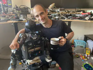 Smiling man leans beside the robot while holding a mug in the electronics-filled workshop.