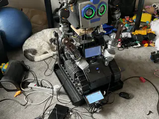 Angled full view of the tracked robot with green ring eyes, connected cables, and a curled dog behind.