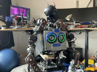 Front upper-body view of the robot's green ring eyes and articulated arms in a cluttered workshop.