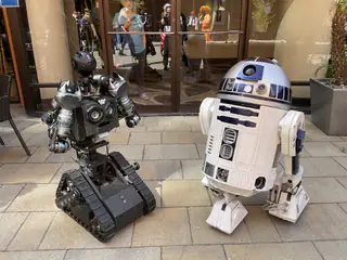 Tracked humanoid robot posed outdoors beside an R2-D2 replica, with people reflected in windows.