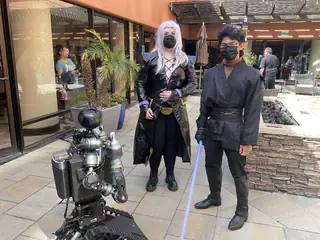 Two masked costumed adults holding illuminated purple and blue prop swords behind the robot outdoors.