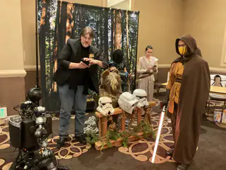 Convention display with costumed adults, a forest backdrop, helmet props, and the tracked robot at left.