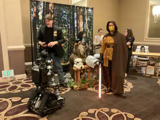 Hooded masked costumed person holding a glowing red prop sword beside the robot and helmet display.