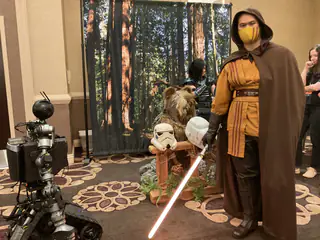 Closer view of a hooded masked costumed person with a red prop sword and forest backdrop.