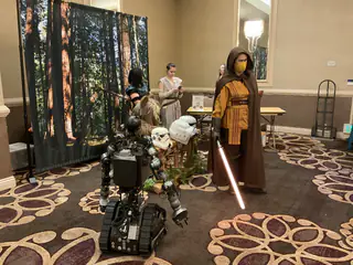 Full-body hooded costumed person holding a glowing red prop sword beside the robot and display helmets.