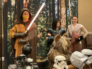Hooded costumed person raises a glowing red prop sword before a group and helmet display.