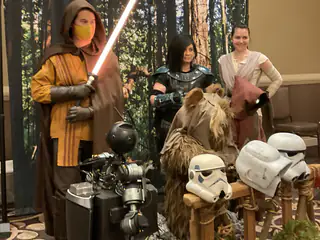 Group of costumed people behind helmet props, with the robot and a raised red prop sword in front.