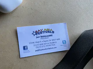 Bay Area Festivals business card lying on beige carpet beside a black strap.