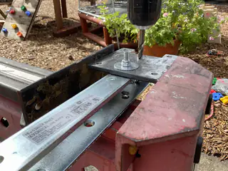 Closer angle of a drill driving a bolt through overlapping metal rails on a red support.