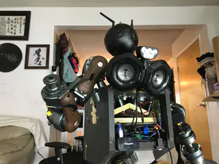 Front upper view of the robot's antennaed spherical head, twin speakers, exposed wiring, and raised arm.
