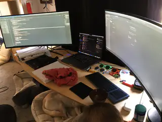 Three-screen coding workstation with a laptop, electronics, and a resting dog beneath the desk.