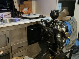 Tracked humanoid robot parked beside kitchen cabinets with blue fabric draped over one shoulder.