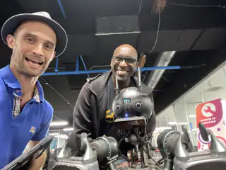 Two smiling men pose behind the robot at an exhibit, with a visitor desk sign nearby.
