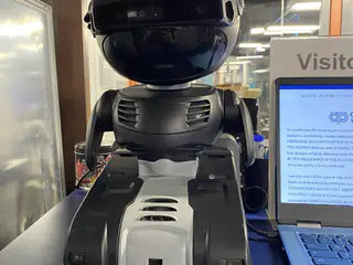 Black and white service robot standing beside a laptop display at a visitor information area.