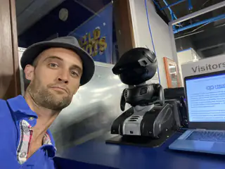 Man in a cap takes a selfie beside a compact service robot and laptop display.