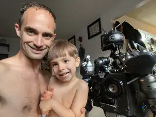 Smiling adult holds a smiling child beside the black humanoid robot in close framing.