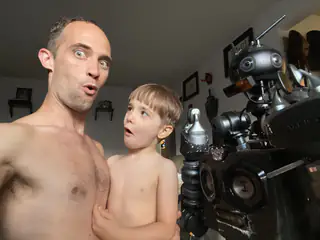 Adult makes a surprised expression while the child looks toward the robot beside them.