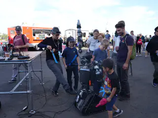 Children and adults gather around a compact tracked robot at an outdoor maker fair.