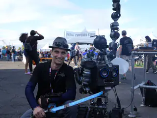 Smiling exhibitor sits beside a tracked robot with round blue eyes and a raised arm.