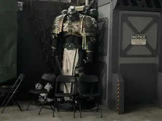 Large armored humanoid robot stands behind empty folding chairs inside a dark industrial exhibit.