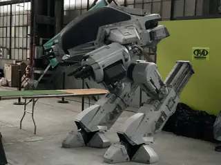 White armored bipedal robot crouches on broad mechanical feet inside an exhibition hall.