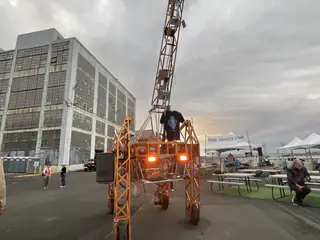 Tall orange open-frame machine faces the camera beside a brick industrial building and crane.