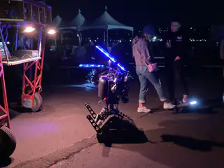 Blue-lit tracked robot approaches two people beneath an orange gantry during a nighttime demonstration.