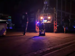 Blue-lit tracked robot sits before an orange gantry as one person stands beside it at night.