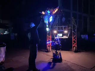 Person stands beside a blue-lit tracked robot under a suspended multicolored dragon head at night.