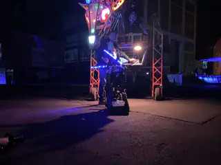 Blue-lit tracked robot faces a lowered multicolored dragon head beneath the orange gantry.