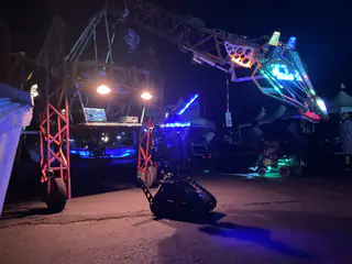 Suspended dragon head stretches sideways over a tracked robot beneath the illuminated gantry.