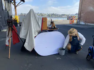 Person in a straw hat kneels beside covered equipment and a low white fabric enclosure.