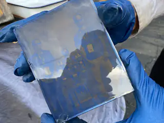 Blue-gloved hands hold a glass plate bearing a faint group portrait around a robot.